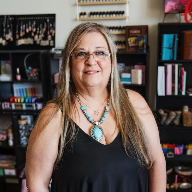 A woman with long hair wearing a black top and turquoise jewelry stands in front of colorful shelves filled with craft supplies.